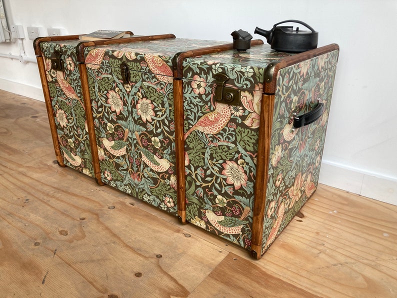 May include: Three vintage wooden storage trunks with a floral and bird patterned design. The trunks are stacked on top of each other and have brass hardware.