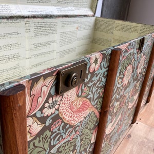 May include: A vintage wooden storage trunk with a floral patterned design. The trunk has a brass lock and hinges. The interior is lined with a cream paper with handwritten text.