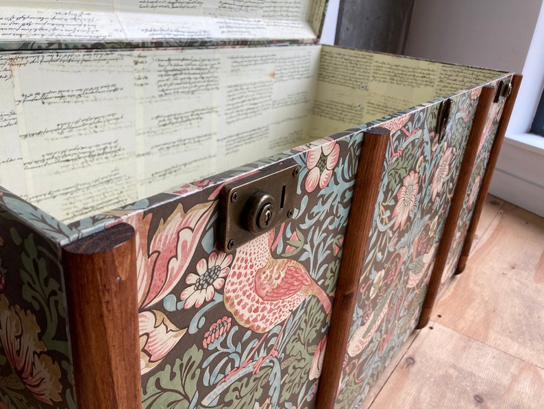 May include: A vintage wooden storage trunk with a floral patterned design. The trunk has a brass lock and hinges. The interior is lined with a cream paper with handwritten text.