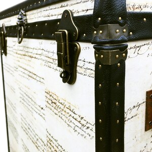 May include: A vintage-style, white leather trunk with black leather trim and brass accents. The trunk has a handwritten inscription on the front and a brass lock and hinge.