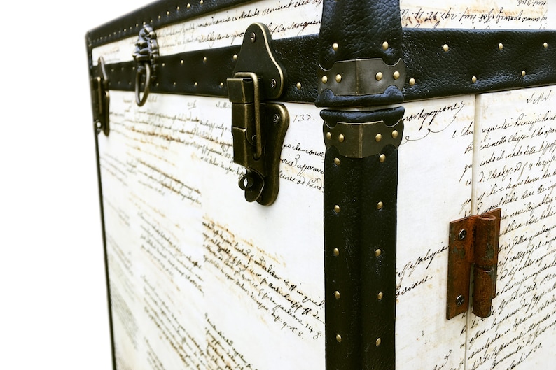 May include: A vintage-style, white leather trunk with black leather trim and brass accents. The trunk has a handwritten inscription on the front and a brass lock and hinge.