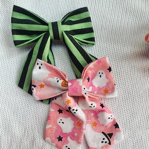 May include: Two fabric hair bows. One bow features green and black stripes. The other bow has a pink background with white ghosts, orange stars, and black stars. The bows are likely for Halloween.