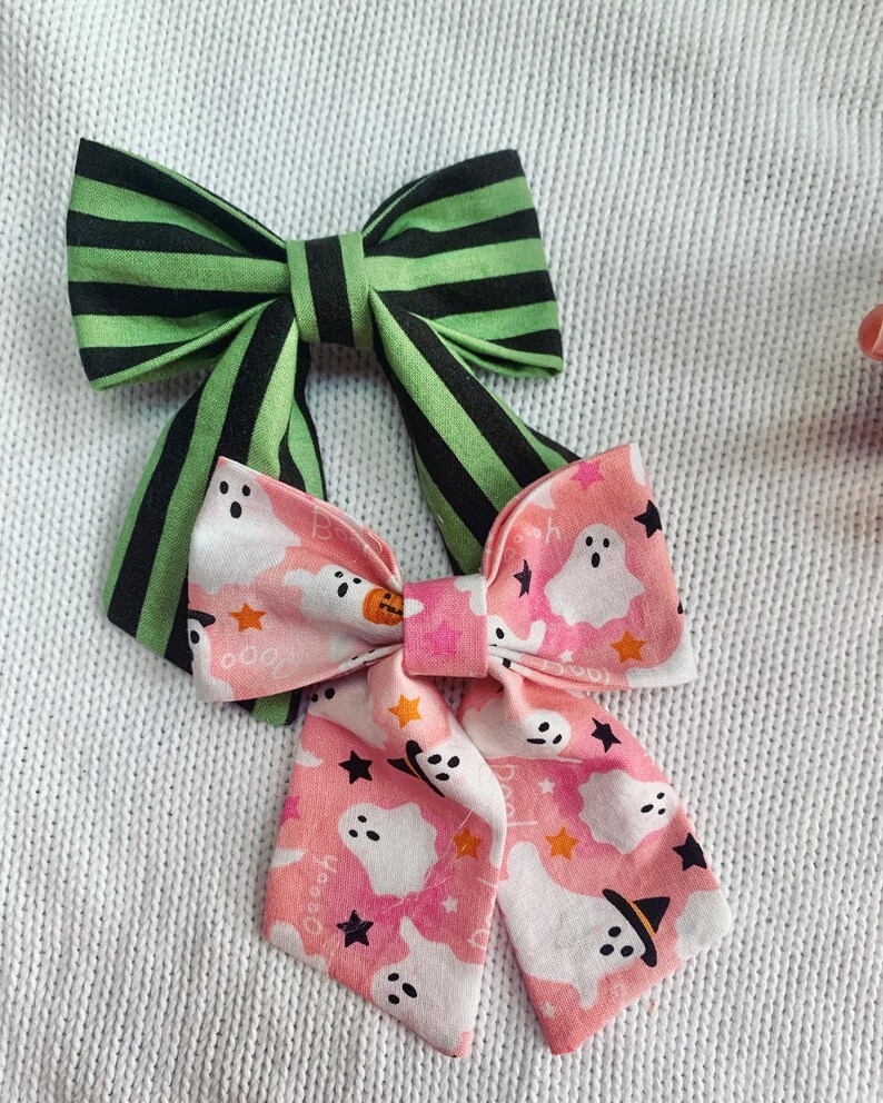 May include: Two fabric hair bows. One bow features green and black stripes. The other bow has a pink background with white ghosts, orange stars, and black stars. The bows are likely for Halloween.