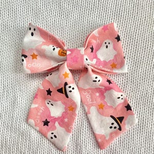 May include: A pink Halloween-themed hair bow with a repeating pattern of white ghosts, black stars, and orange stars. The bow features the word "Boo!" and a pumpkin graphic. The bow is made of fabric.
