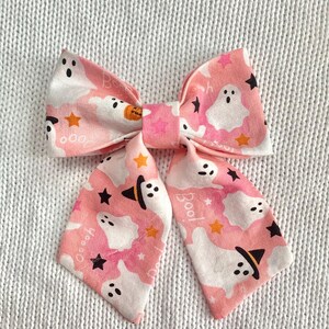 May include: A pink Halloween-themed hair bow with a repeating pattern of white ghosts, black stars, and orange stars. The bow features the word "Boo!" and a pumpkin graphic. The bow is made of fabric.