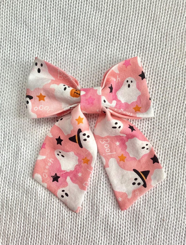 May include: A pink Halloween-themed hair bow with a repeating pattern of white ghosts, black stars, and orange stars. The bow features the word "Boo!" and a pumpkin graphic. The bow is made of fabric.
