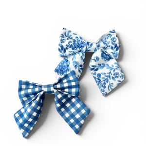 Könnte beinhalten: Zwei Haarschleifen aus Stoff. Eine Schleife zeigt ein blau-weißes Vichy-Karomuster. Die zweite Schleife hat ein blau-weißes Blumenmuster mit gemalten Blättern und Blüten auf weißem Grund. Beide Schleifen haben eine klassische Form.