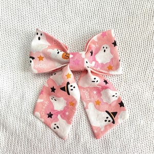May include: A pink Halloween-themed bow tie with a repeating pattern of white ghosts, orange stars, and black witch hats. The bow tie has the word "Boo!" printed on the tails. The bow tie is made of fabric.