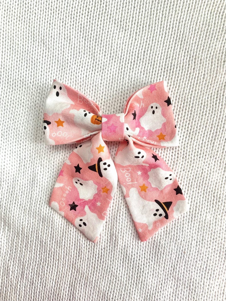 May include: A pink Halloween-themed bow tie with a repeating pattern of white ghosts, orange stars, and black witch hats. The bow tie has the word "Boo!" printed on the tails. The bow tie is made of fabric.