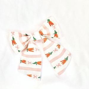 May include: A white and pink striped hair bow with a carrot print. The bow has a pink ribbon tied in the center.
