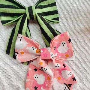 May include: Two handmade Halloween-themed hair bows. One bow features green and black stripes, while the other is pink with white ghosts, black stars, and orange pumpkins. The bows are made of fabric and are likely for children.