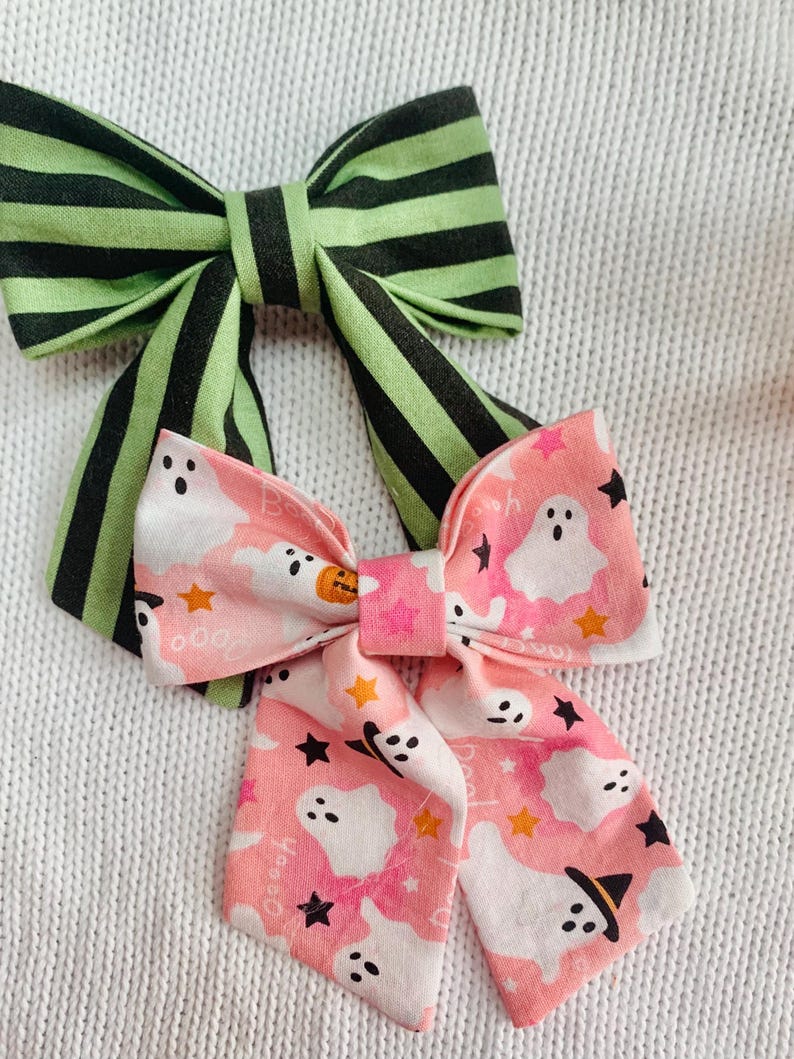 May include: Two handmade Halloween-themed hair bows. One bow features green and black stripes, while the other is pink with white ghosts, black stars, and orange pumpkins. The bows are made of fabric and are likely for children.