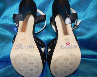 May include: A pair of black high-heeled sandals with rhinestones spelling "Team" on one sole and "Bride" with a pink heart on the other.