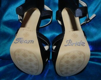 May include: A pair of black high-heeled sandals with a rhinestone embellishment on the sole. One sandal says "Team" and the other says "Bride".