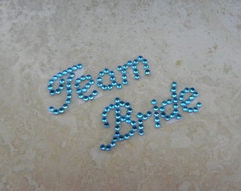 May include: Two separate phrases, "Team" and "Bride", spelled out in sparkling blue rhinestones. The rhinestones are arranged to form cursive lettering. The background is a light, textured surface.