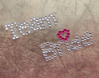 May include: Team Bride rhinestone iron-on patch with a pink heart in the center. The patch is made of clear adhesive backed rhinestones.