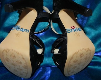 May include: A pair of black high-heeled sandals with a decorative pattern on the soles. One sandal has "Team" written on the sole in blue rhinestones, and the other has "Bride" written on the sole in blue rhinestones with a red heart.