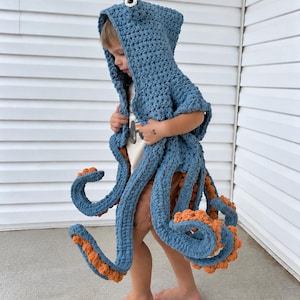 May include: A blue crocheted octopus hooded cape featuring long dangling tentacles with textured orange undersides and a single prominent eye detail on the top of the hood. The garment is crafted from thick yarn with a visible stitch pattern.