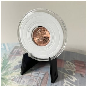 Puede incluir: Un centavo de cobre con la fecha 5-17 y las iniciales "S & C" grabadas en él, dentro de un portamonedas de plástico transparente con fondo blanco, sobre un soporte negro.