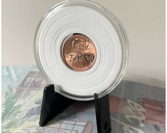 May include: A copper penny with the date 5-17 and the initials "S & C" engraved on it, inside a clear plastic coin holder with a white background, on a black stand.