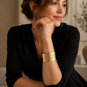 Può includere: Un ampio bracciale a fascia color oro opaco con testo inciso in corsivo. Il cinturino in metallo ha una finitura liscia e spazzolata e avvolge il polso, mostrando una chiara iscrizione di colore scuro incisa sulla superficie del metallo.