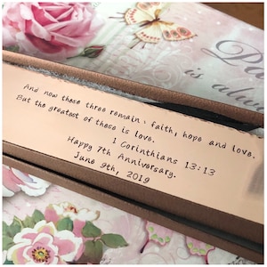 May include: A close-up of a brown leather box with a quote from 1 Corinthians 13:13 inscribed on the top. The quote reads "And now these three remain: faith, hope and love. But the greatest of these is love." The box also includes the date "June 9th, 2019" and the words "Happy 7th Anniversary."