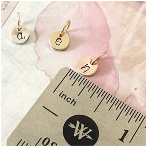Puede incluir: Tres peque&ntilde;os dijes circulares de metal con las letras min&uacute;sculas a, e y n. Los dijes est&aacute;n colocados junto a una regla de metal con medidas en pulgadas para indicar su peque&ntilde;o tama&ntilde;o en relaci&oacute;n con la escala.