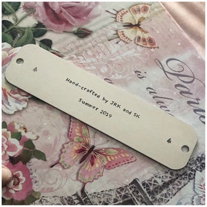 Peut inclure: Un marque-page en argent avec le texte "Hand-crafted by JRK and SK Summer 2019" gravé dessus. Le marque-page est sur un fond floral rose et blanc.