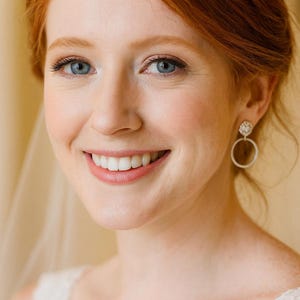 May include: A woman with red hair and blue eyes smiles, wearing a white lace top and hoop earrings. The image is a close-up portrait, with a soft focus on the woman's face. The background is a light, neutral colour.
