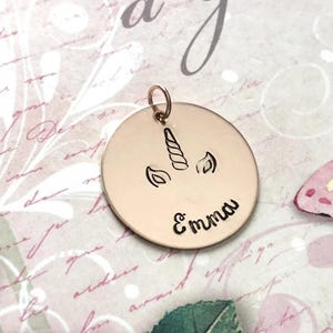 May include: A circular rose gold pendant featuring an engraved unicorn horn and ears design centered above the name Emma in a cursive script. The pendant includes a small circular jump ring at the top for attachment to a chain or bracelet.