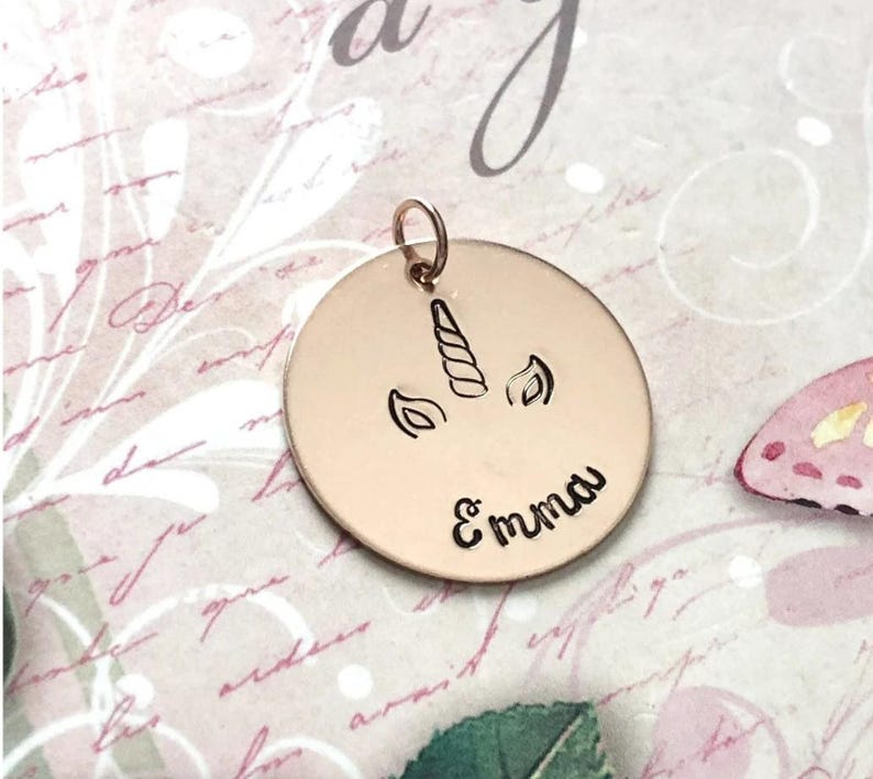 May include: A circular rose gold pendant featuring an engraved unicorn horn and ears design centered above the name Emma in a cursive script. The pendant includes a small circular jump ring at the top for attachment to a chain or bracelet.