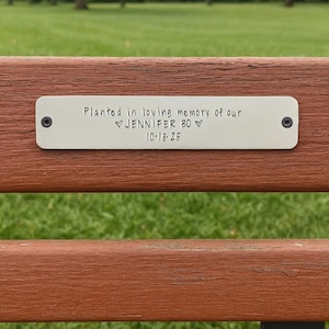 May include: A close-up of a wooden park bench with a metal plaque. The plaque reads "Planted in loving memory of our Jennifer BO 10.18.25". The bench is brown with a green grassy background and trees.