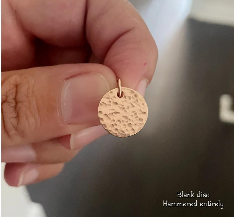May include: A small, circular rose gold pendant featuring a textured, hammered finish across its entire surface. The disc includes a small jump ring at the top for attachment. The text "Blank disc Hammered entirely" is printed in the corner.