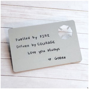 May include: A silver metal card with a cut-out firefighter's symbol. The card has the text "Fuelled by FIRE Driven by COURAGE Love you always Goose" written on it.