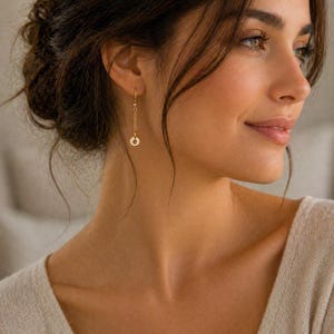 May include: Gold-colored dangle earrings with a small circular charm featuring the letter "e". The earrings hang from a delicate chain and are worn on the ear. The earrings are a simple yet elegant accessory.