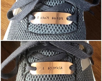 May include: A pair of metallic copper-colored shoe lace tags threaded onto grey mesh athletic sneakers. The top tag is engraved with the text I don't sweat, and the bottom tag is engraved with the text I sparkle, both in a simple sans-serif font.