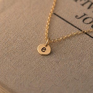 May include: A gold-toned necklace featuring a delicate chain and a small, circular pendant. The pendant is engraved with the lowercase letter e in a simple, black, sans-serif font. The chain is composed of small, interconnected oval links.