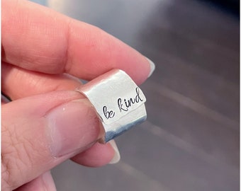 May include: A silver ring with the words "be kind" engraved on it.