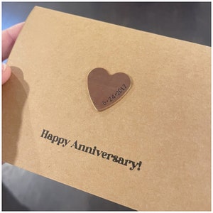Puede incluir: Una tarjeta marr&oacute;n con el texto "Happy Anniversary!" y un coraz&oacute;n de cobre con la fecha "6-24-2017" grabada en &eacute;l.