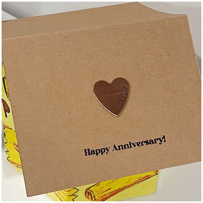 Puede incluir: Una tarjeta marr&oacute;n con el texto "Happy Anniversary!" impreso en ella. Un peque&ntilde;o amuleto de coraz&oacute;n dorado con la fecha "9-24-2017" grabada en &eacute;l est&aacute; unido a la tarjeta.