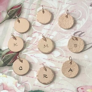 May include: A collection of nine circular rose gold metal charms, some with smooth surfaces and others with a hammered texture. Several charms are stamped with individual uppercase letters including K, M, E, C, R, and A, each featuring a small jump ring.