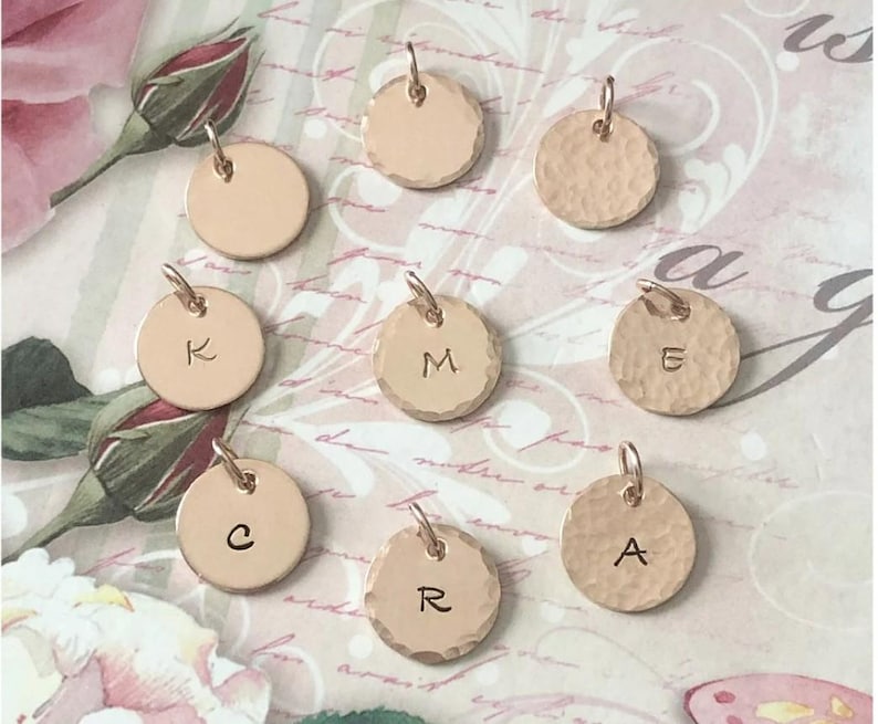 May include: A collection of nine circular rose gold metal charms, some with smooth surfaces and others with a hammered texture. Several charms are stamped with individual uppercase letters including K, M, E, C, R, and A, each featuring a small jump ring.