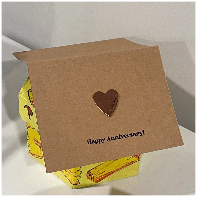 Puede incluir: Una tarjeta marr&oacute;n con las palabras "Happy Anniversary!" impresas en ella. Un peque&ntilde;o coraz&oacute;n de oro con la fecha "10.28.2017" grabada en &eacute;l est&aacute; unido a la tarjeta.