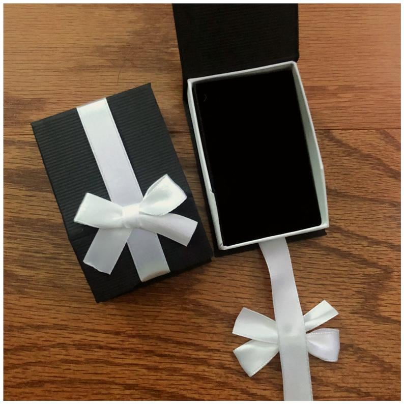 May include: A black gift box with a white ribbon and bow. The box is open and the lid is laying on the table. The ribbon is tied into a bow on the lid of the box. The box is on a wooden surface.