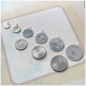 May include: A collection of silver charm pendants with various designs, including a heart, a letter J, a sunburst, the words "Faith Over Fear", and a footprint with the name "Leo".
