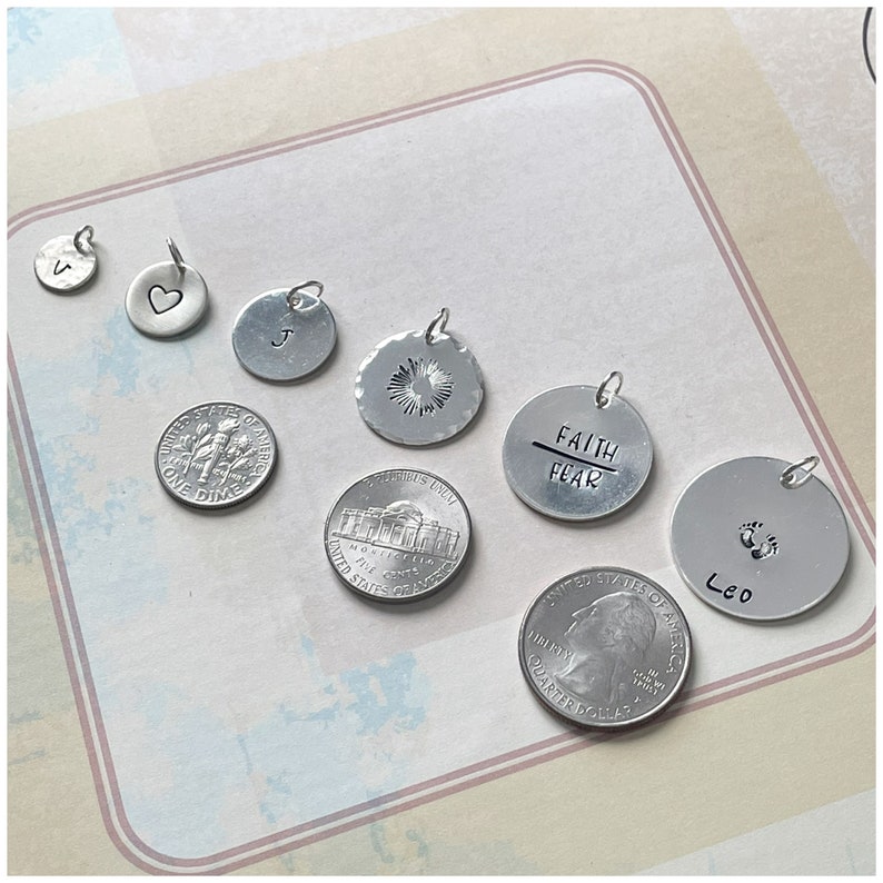 May include: A collection of silver charm pendants with various designs, including a heart, a letter J, a sunburst, the words "Faith Over Fear", and a footprint with the name "Leo".