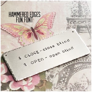 May include: A rectangular silver-toned metal tag with hammered edges and two small holes at the ends. The tag features stamped text reading "CLOSE - close blind" with a downward arrow and "OPEN - open blind" with an upward arrow in a rounded font.