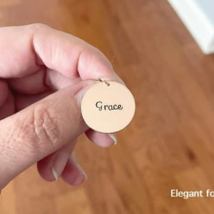 May include: A circular gold-toned metal pendant featuring the name Grace engraved in a dark script font. The pendant includes a small jump ring at the top for attachment. The text Elegant font is visible in the lower right corner of the image.