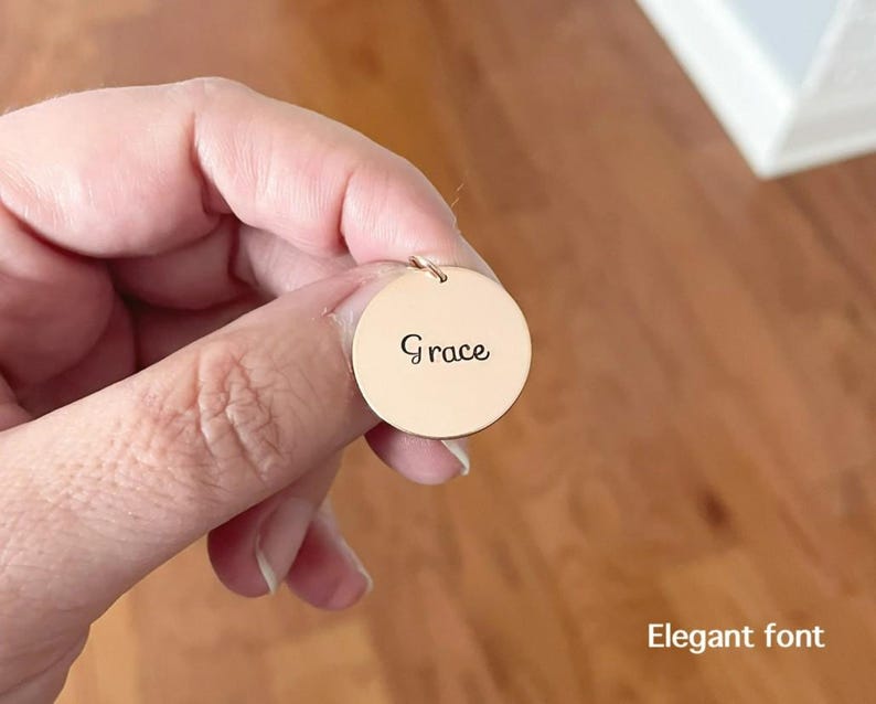 May include: A circular gold-toned metal pendant featuring the name Grace engraved in a dark script font. The pendant includes a small jump ring at the top for attachment. The text Elegant font is visible in the lower right corner of the image.