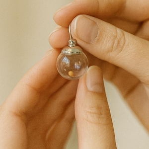 May include: A clear glass earring with a small, round, silver-coloured top and a tiny, round, yellow object inside. The earring is held between two hands against a neutral background.